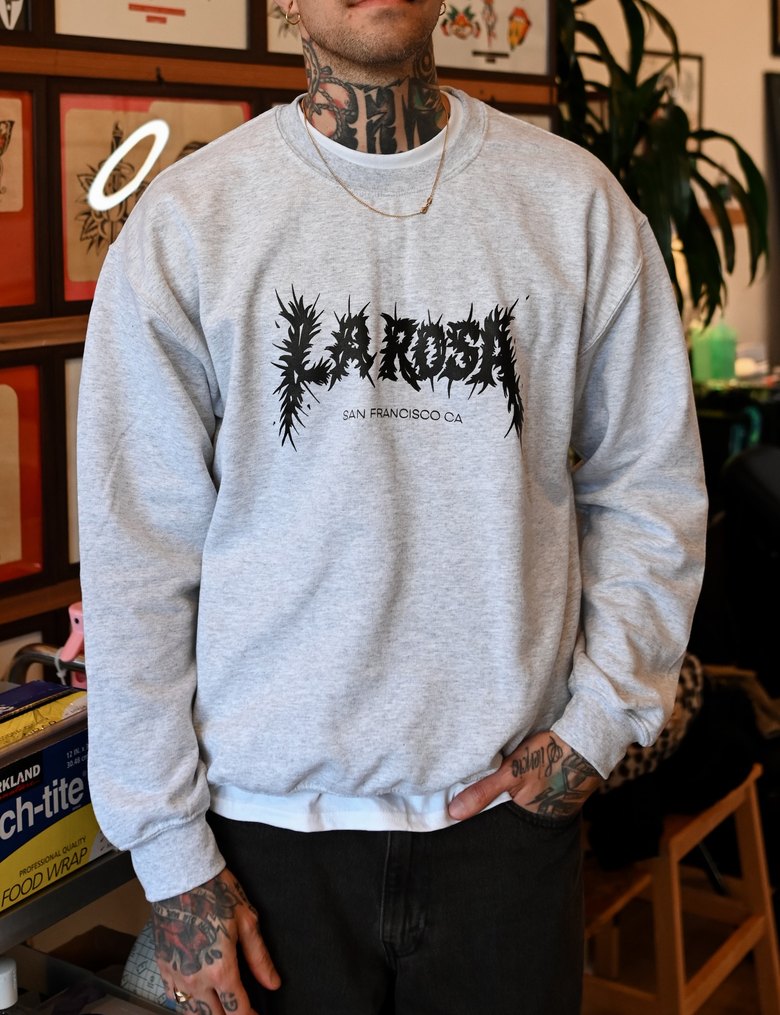 A grey La Rosa San Francisco crewneck worn in the shop, a neon ring glowing on the wall behind.