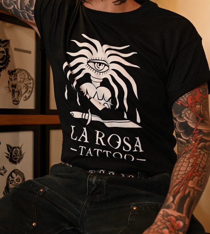 A black La Rosa Tattoo tee with a white eye, heart and dagger print, worn seated in the shop.