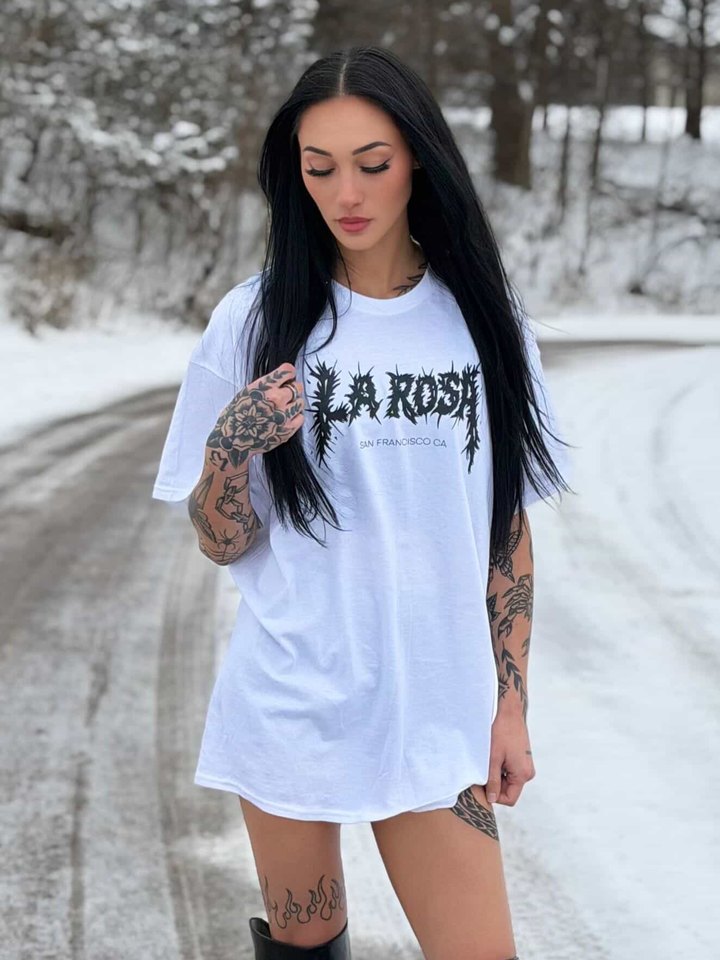 An oversized white La Rosa tee worn as a dress on a snowy road, sleeves of tattoos showing.