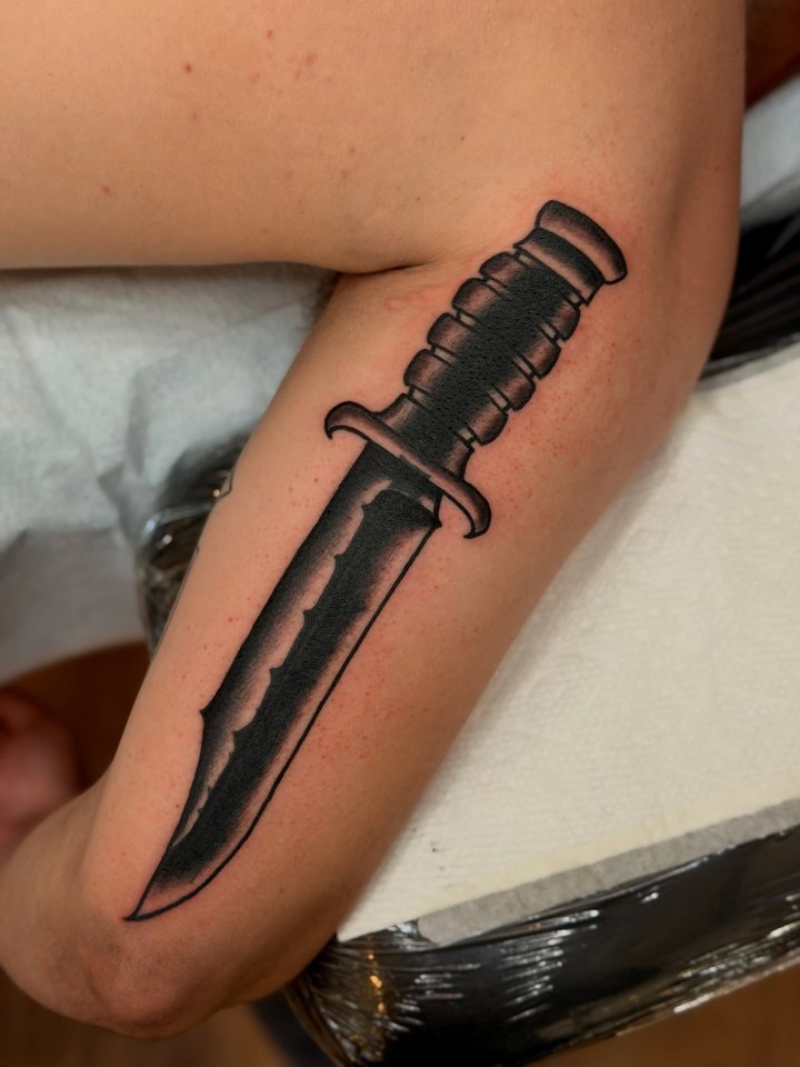 A black and grey traditional combat dagger tattooed along a forearm.