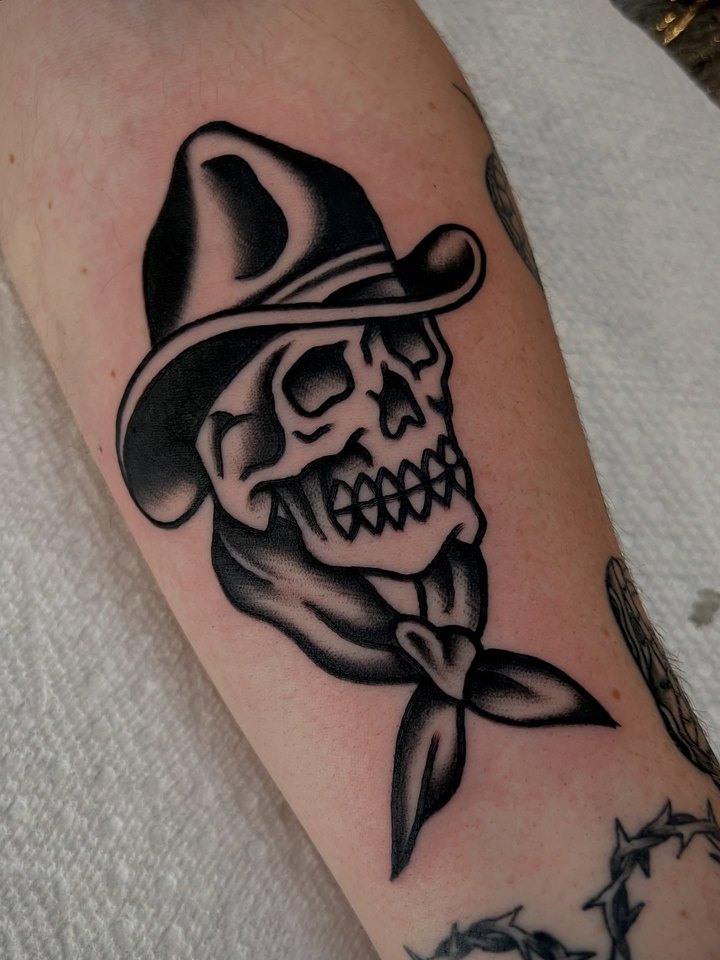 A bold black traditional cowboy skull with hat and bandana tattooed on a forearm.