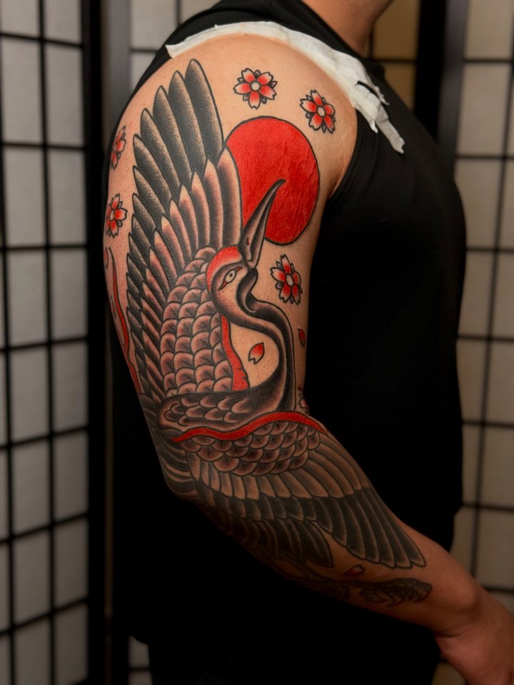 A Japanese crane with a red rising sun and cherry blossoms covering an upper arm.