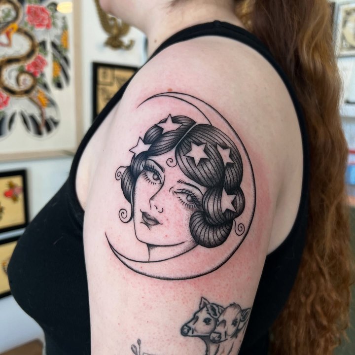 A crescent moon with a woman's face tattooed in black and grey on a shoulder.