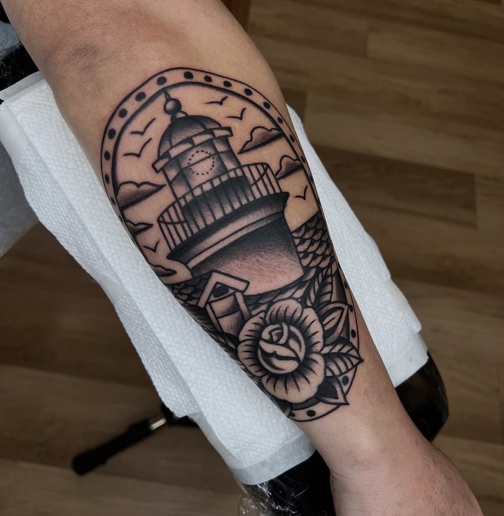 An American traditional lighthouse with a rose and waves in an arched frame, on a forearm.