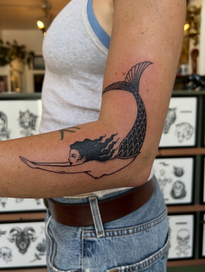 A black and grey mermaid tattoo wrapping an upper arm, shop flash frames visible behind.