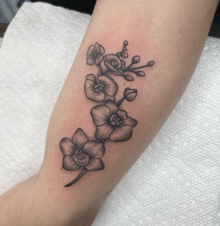 A fine-line black and grey orchid branch tattooed along the inside of a forearm.