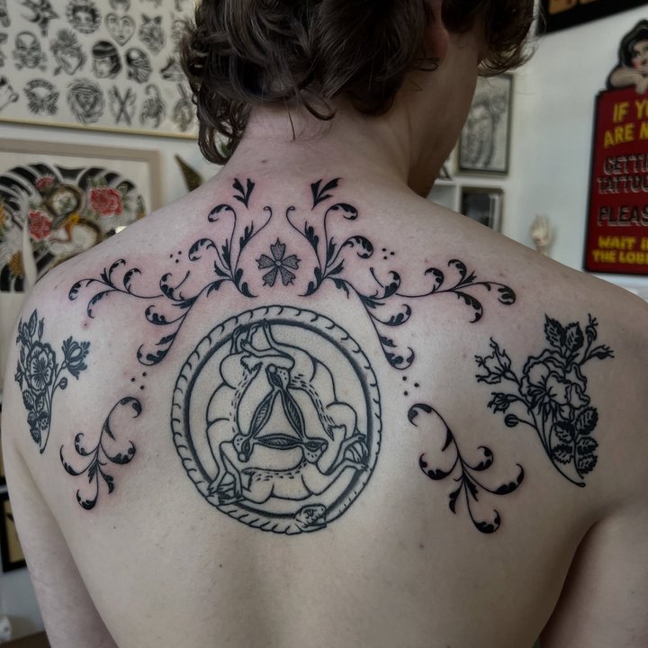 Ornamental blackwork filigree with a three-hares medallion and flowers tattooed across an upper back.