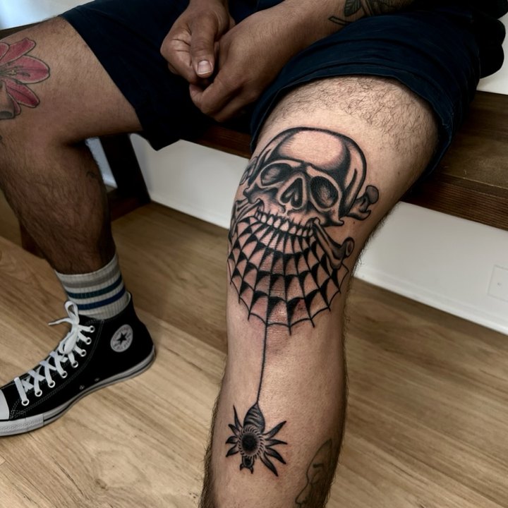 A skull and crossbones over a draped spiderweb tattooed on a knee, a spider hanging below on a thread