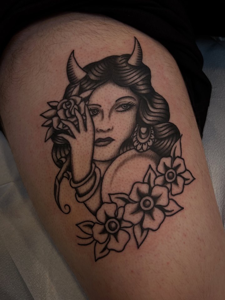 A black and grey lady head with devil horns and flowers, tattooed on a thigh