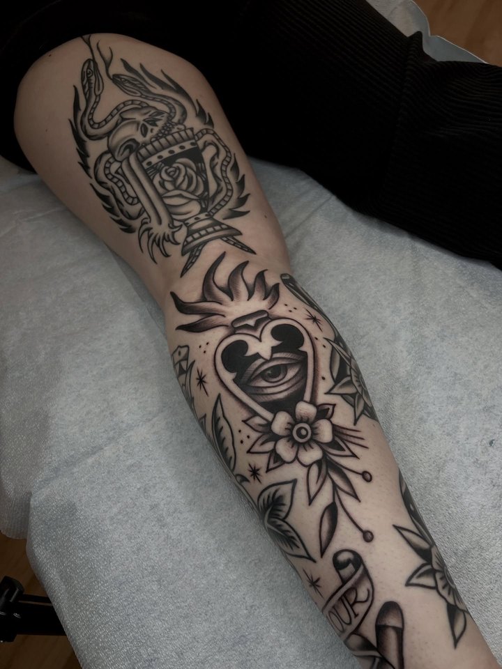 A black and grey leg sleeve: a winged skull urn pierced by a dagger at the thigh, a flaming sacred heart with an eye over flowers on the calf