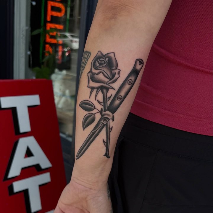 A black and grey rose and switchblade on a forearm, photographed outside the shop.